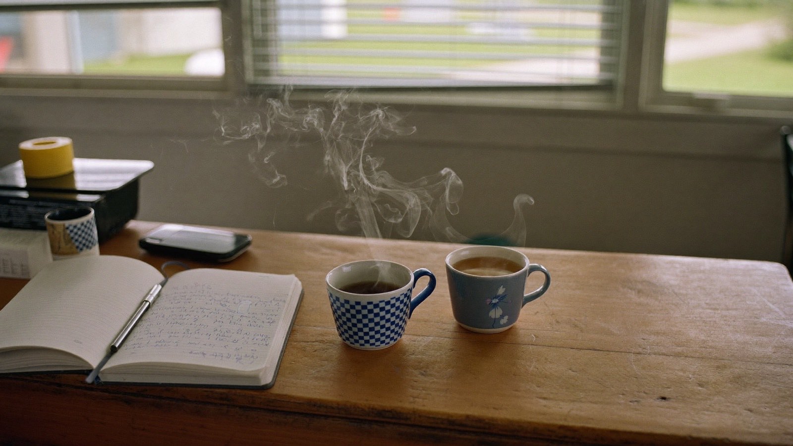 Two mugs of tea and an open notebook with pen on a worn timber kitchen table, morning light through window blinds.