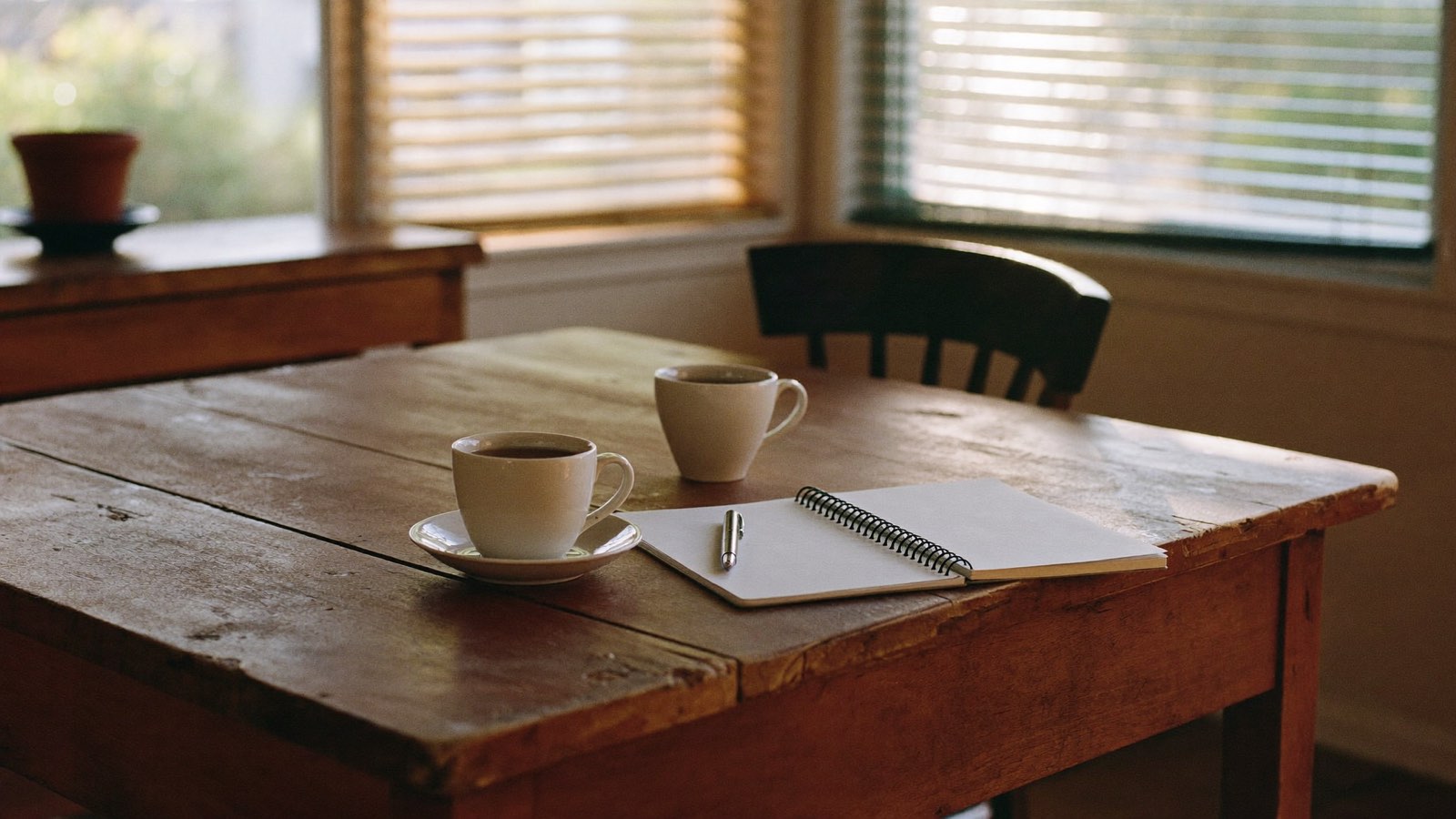 Editorial photograph of two mugs and an open notebook on a worn timber kitchen table in morning light.