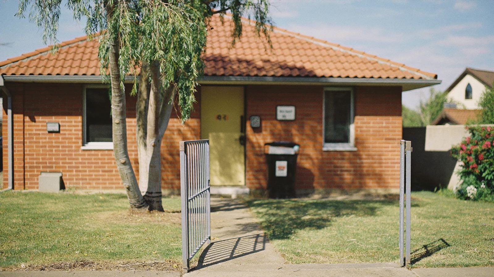 A red-brick suburban Sydney house with a terracotta tiled roof, its front gate standing open and a gum tree shading the lawn