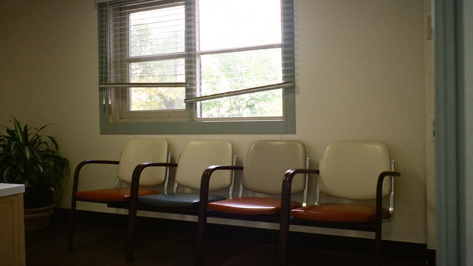 Photograph of four empty vinyl chairs against a plain wall in a small clinic waiting area, daylight through venetian blinds.