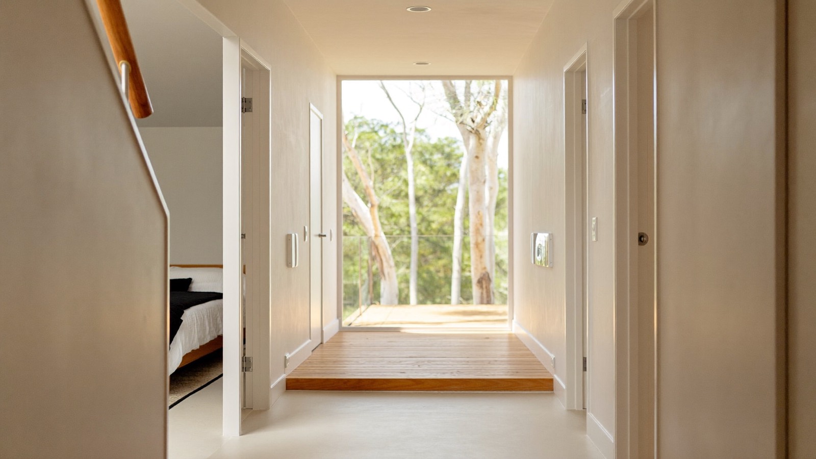 A wide bright hallway in a modern single-storey home, a bedroom off one side and a full-height glazed opening onto a deck with gum trees.