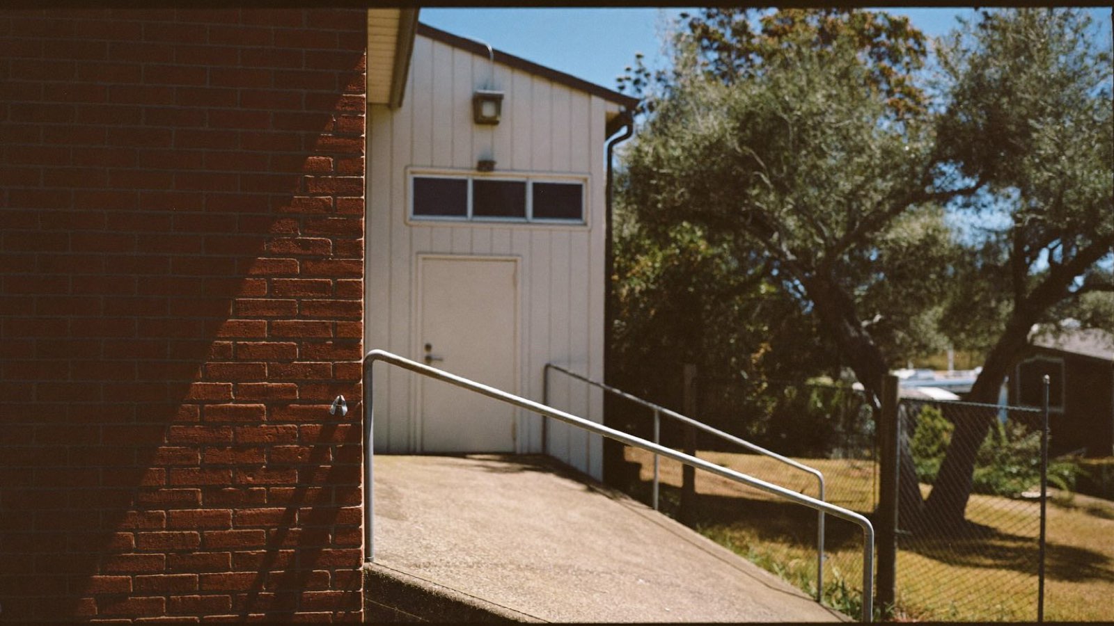 A concrete access ramp with a steel handrail leading to the side door of a brick and weatherboard suburban hall in bright daylight.