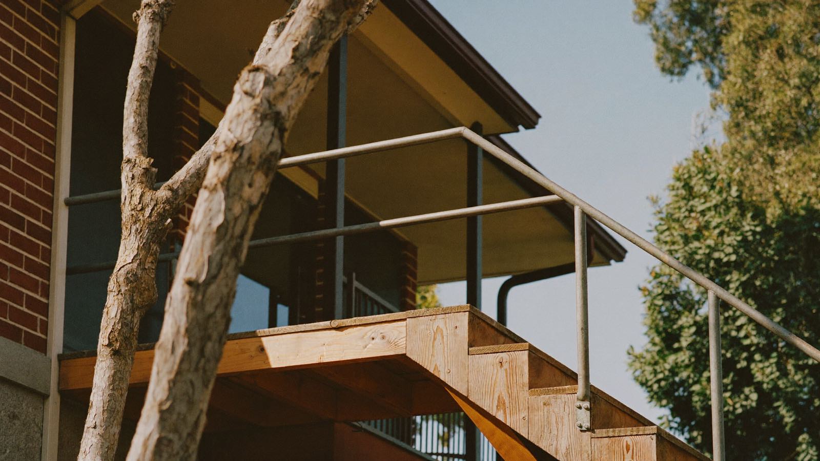 Editorial photograph of a timber landing and steps with a metal handrail against a red-brick suburban home, a gum tree in the foreground.