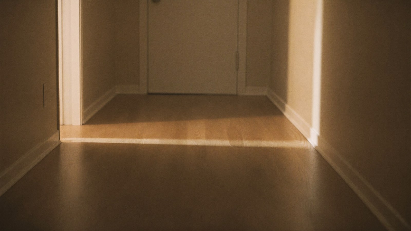Photograph of an empty suburban hallway at night, warm light spilling across timber floorboards from an open doorway.