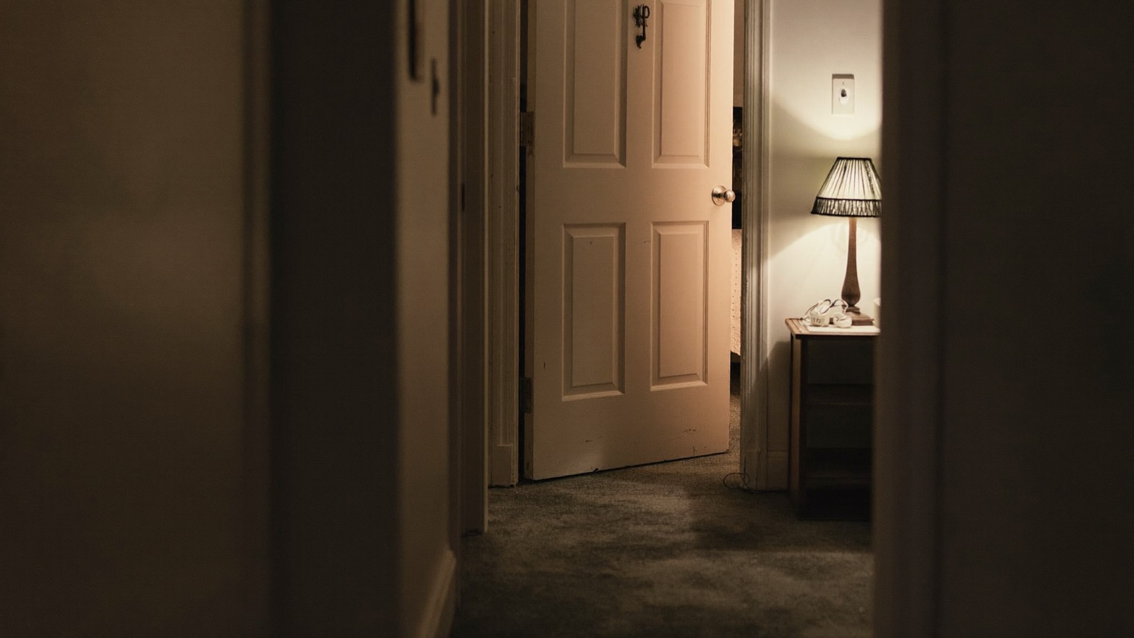 A dimly lit suburban hallway at night with a bedroom door ajar and a lamp glowing on a side table.