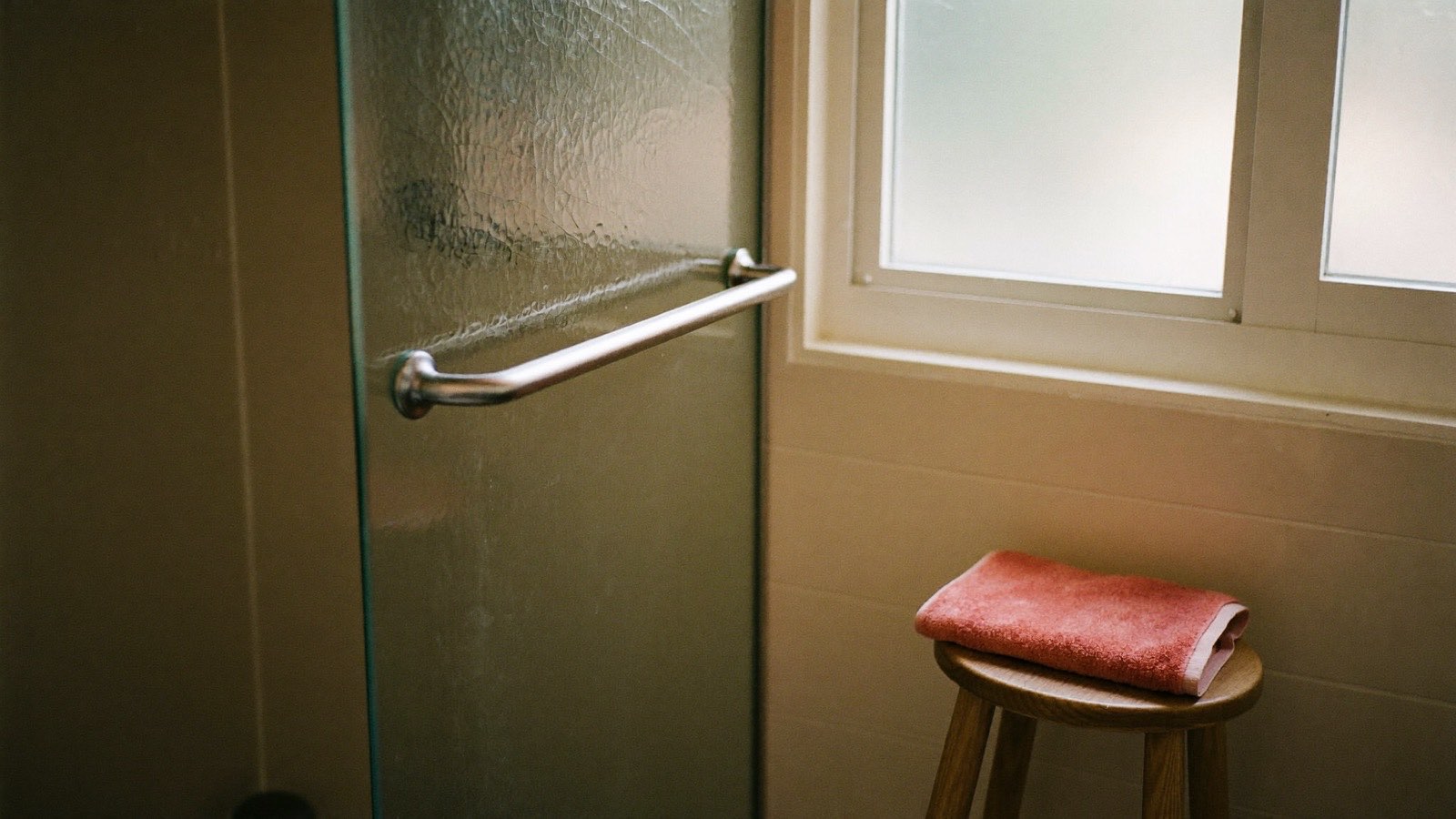 A domestic bathroom with a grab rail beside the shower, a folded towel on a timber stool and morning light through frosted glass.