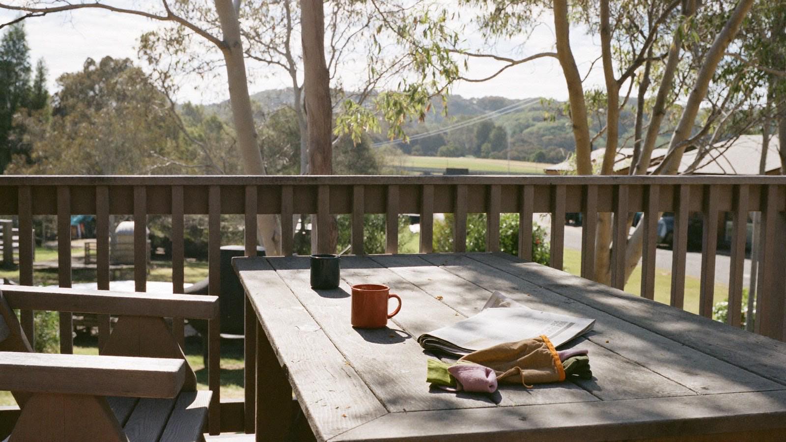 A suburban back deck on a bright morning, with two mugs, a folded newspaper and gardening gloves on a weathered timber table.