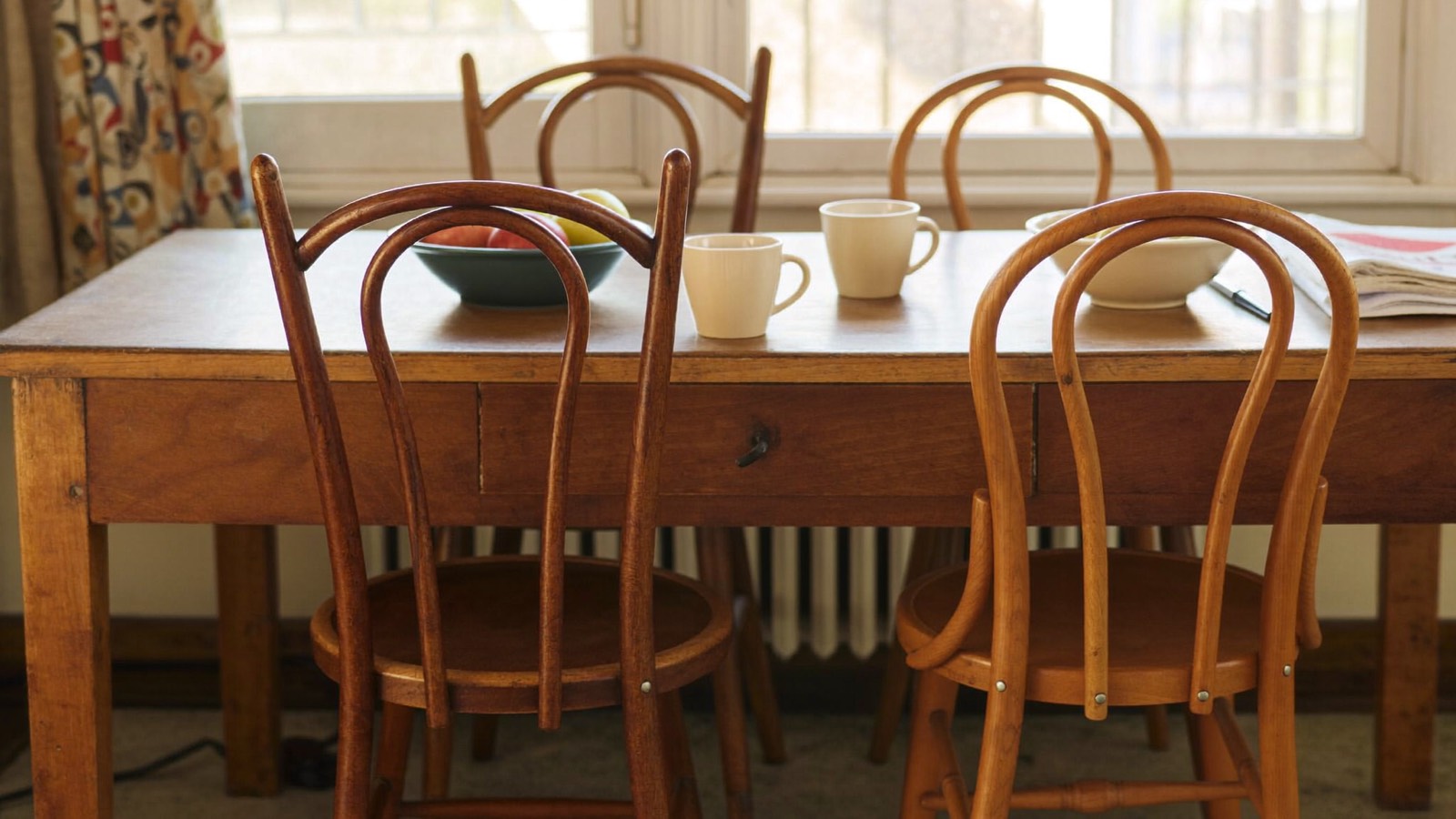 Four mismatched bentwood chairs pulled up to a worn timber table in a share home, plain mugs and a bowl of apples on top