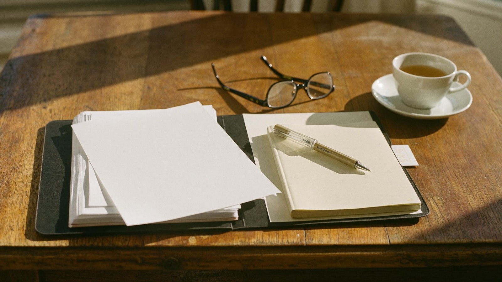 A timber table in afternoon light with an open folder of blank paper, a notebook, a pen, reading glasses and a cup of tea