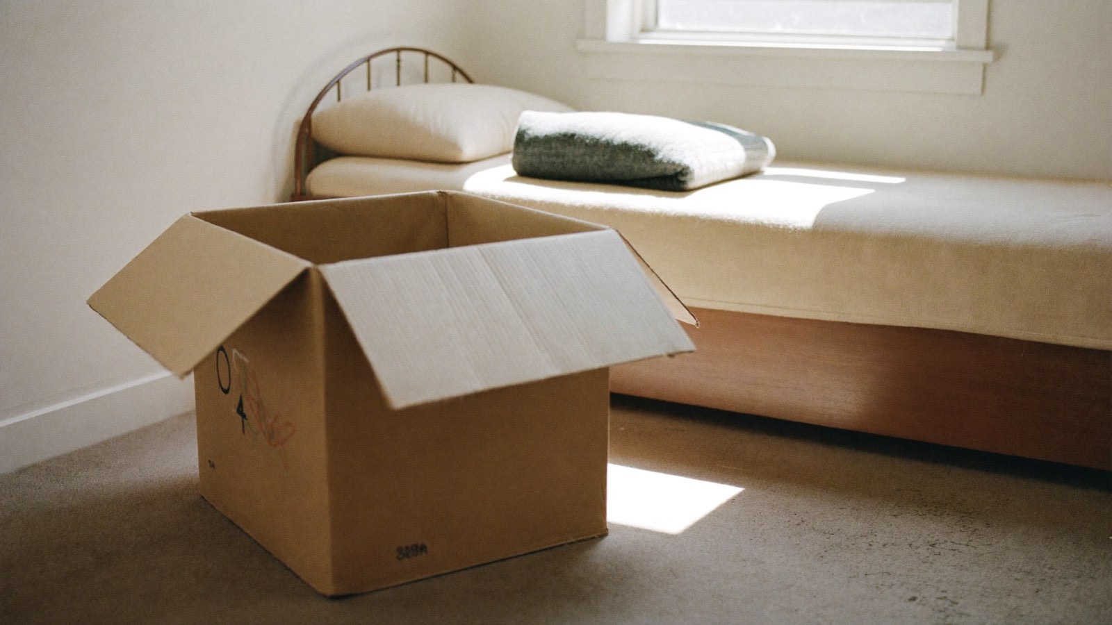 Photograph of an open cardboard moving box on the carpet of a plain bedroom, beside a neatly made single bed in morning light.