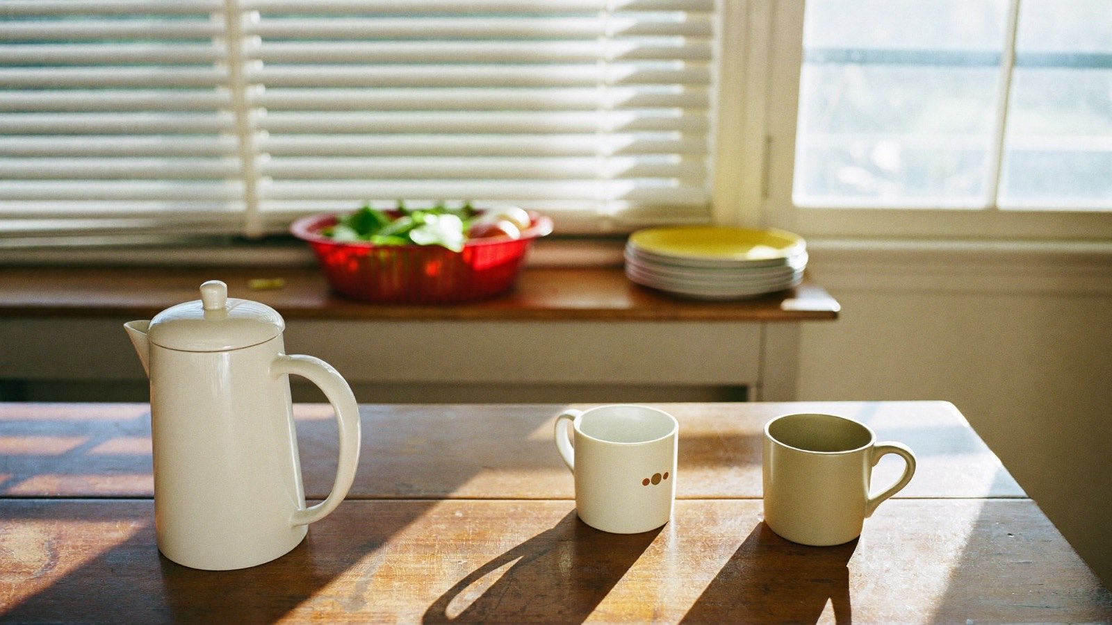 A sunlit shared-home kitchen in suburban Sydney with a pot of tea and two mugs on a worn timber table, morning light through venetian blinds