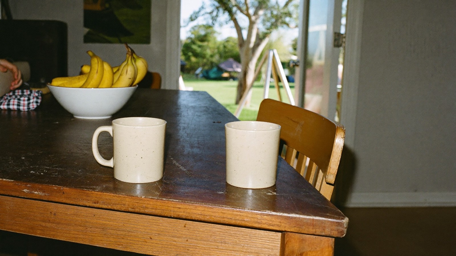 Two plain cream mugs and a bowl of bananas on a scratched timber table, a back door open onto a sunlit yard with a gum tree.