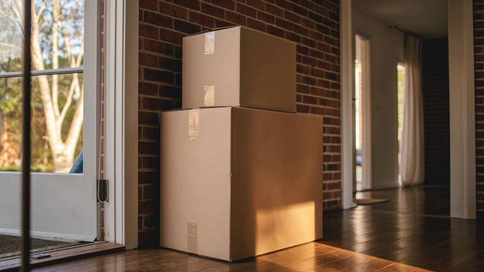 Two plain cardboard moving boxes stacked against a red-brick wall beside an open front door, afternoon light on the floorboards.