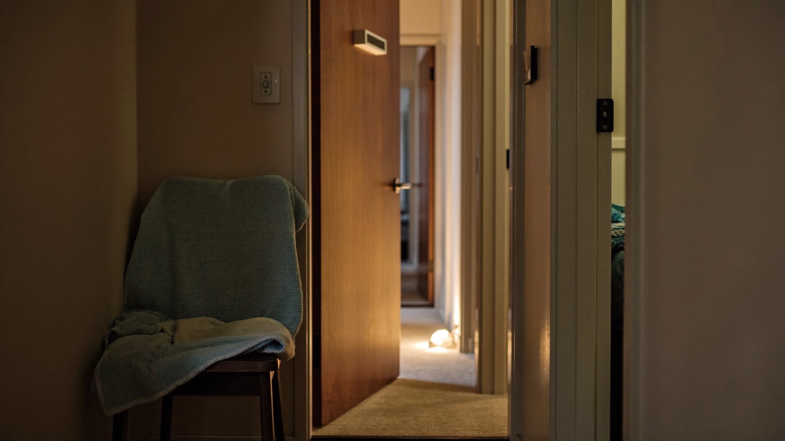The hallway of a share house at night, one bedroom door ajar with warm lamplight spilling onto the carpet, a blanket over a chair.