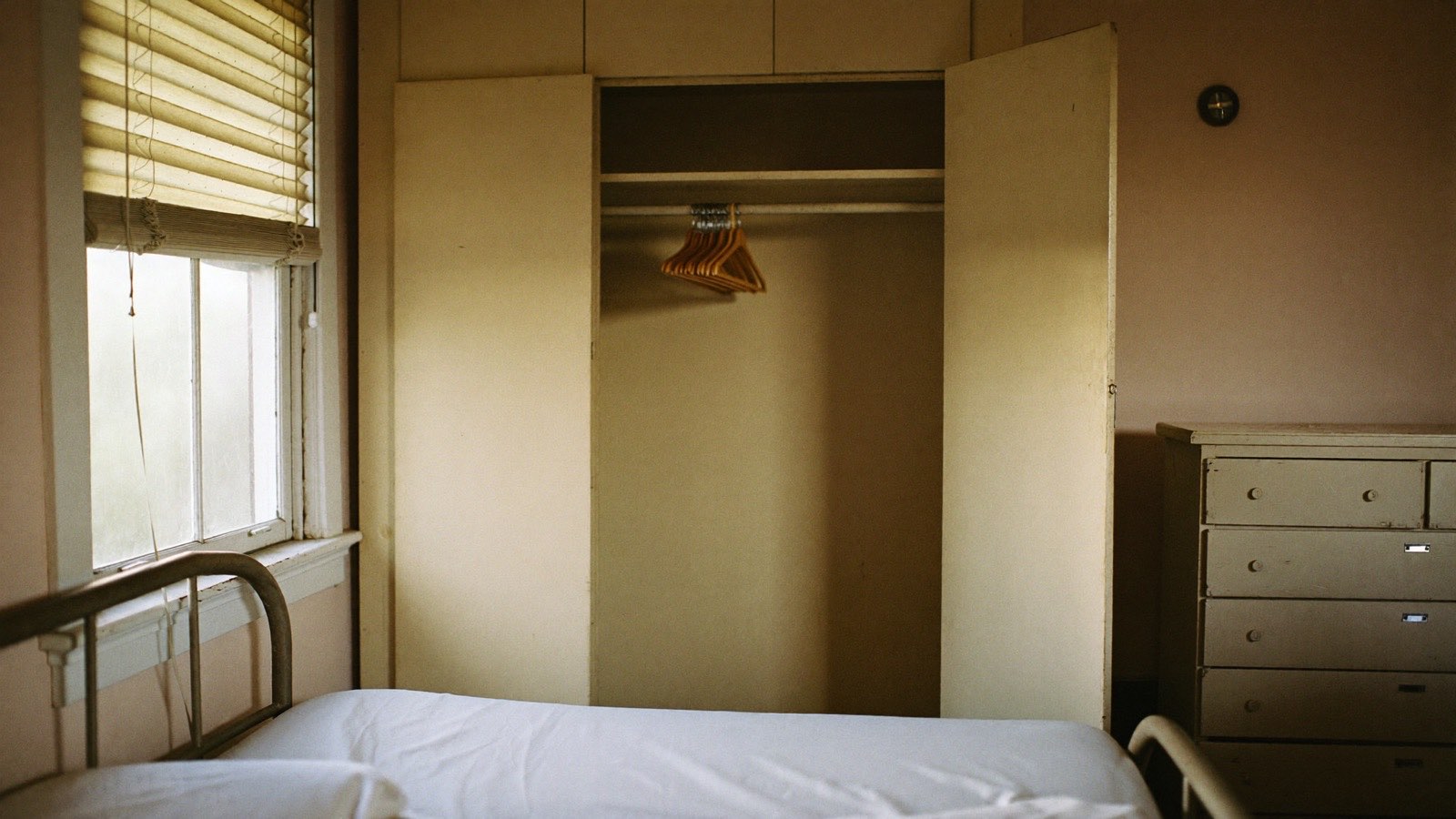 An empty bedroom in an older suburban house, a made-up single bed by the window, a built-in wardrobe open with wooden hangers.