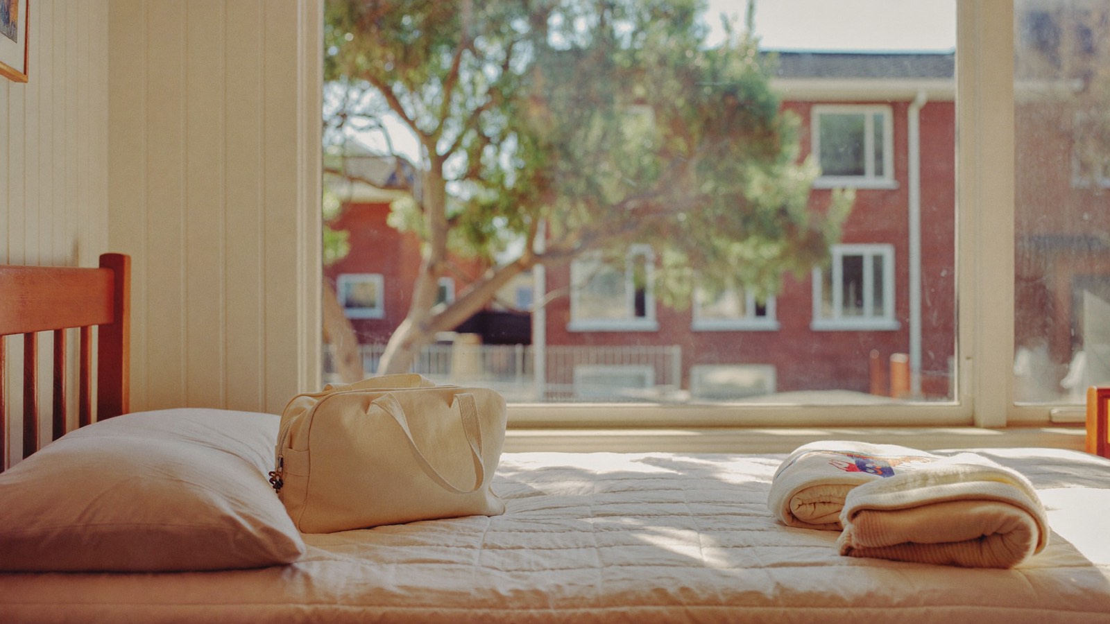 A made single bed with a canvas overnight bag and a folded blanket, a window behind it looking onto a gum tree and a red-brick house.