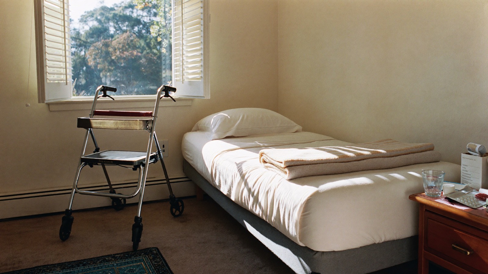 A bright suburban bedroom with a neatly made single bed, a wheeled walking frame parked beside it and a glass of water on the bedside table.
