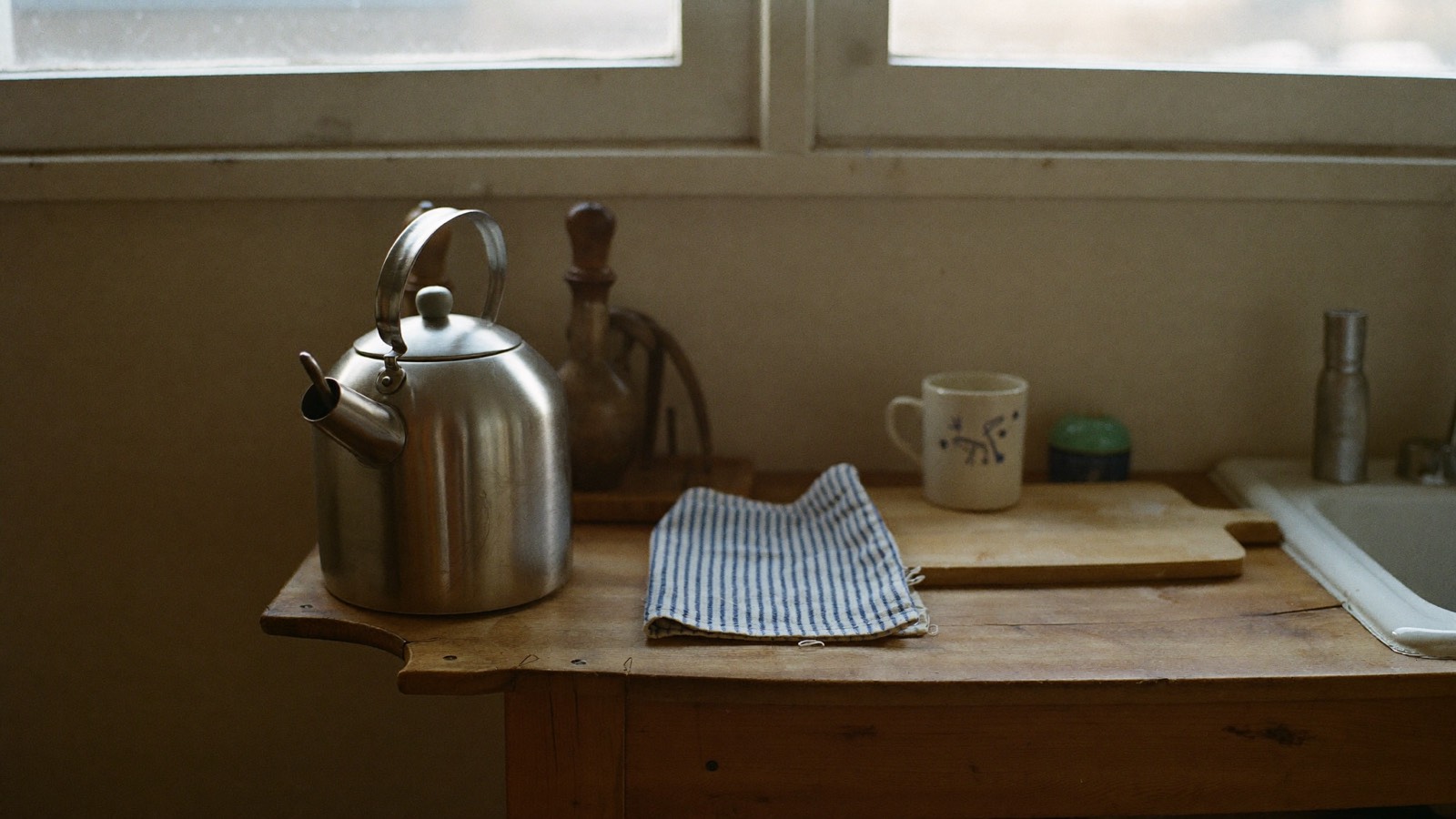 A stainless steel kettle, a folded striped tea towel, a chopping board and a plain mug on a timber kitchen bench beside a window