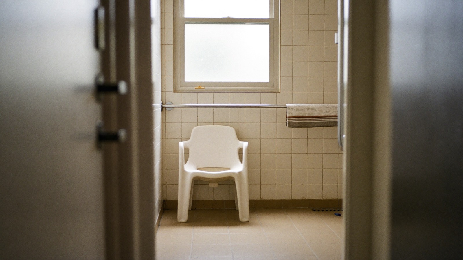 A tiled suburban bathroom seen from the hallway, with a shower chair, a long grab rail and a towel folded over it.