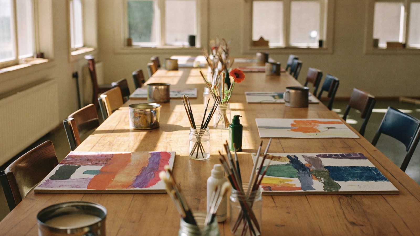 Sunlit community centre art room with paint pots, brushes and unfinished artworks spread across timber tables
