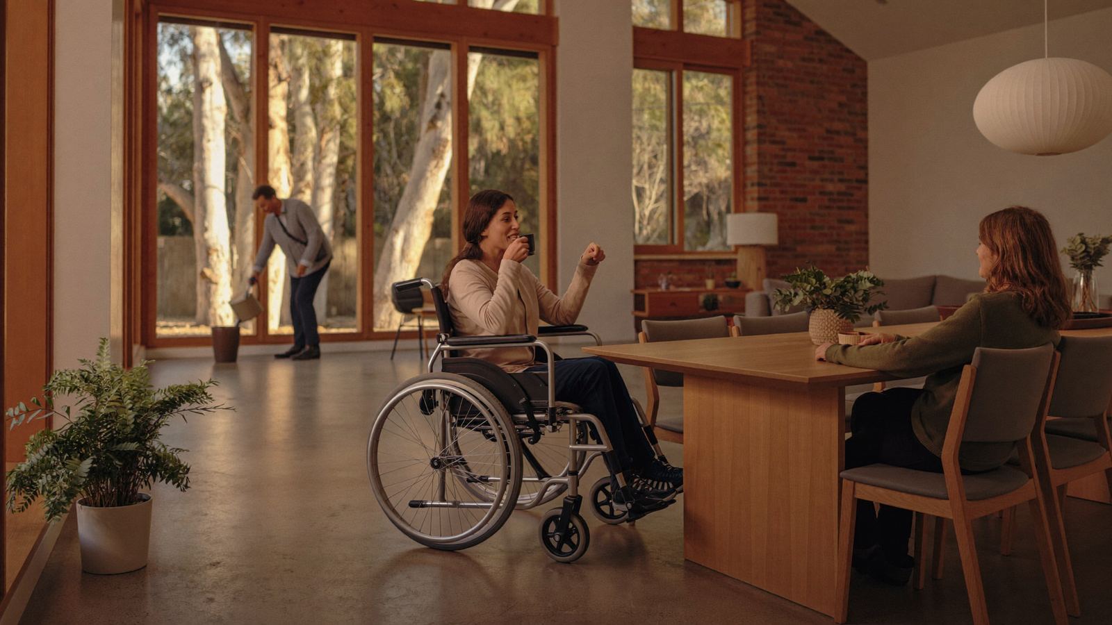 Woman in a wheelchair sharing a cup of tea at the dining table with a support worker while a housemate waters plants by the window